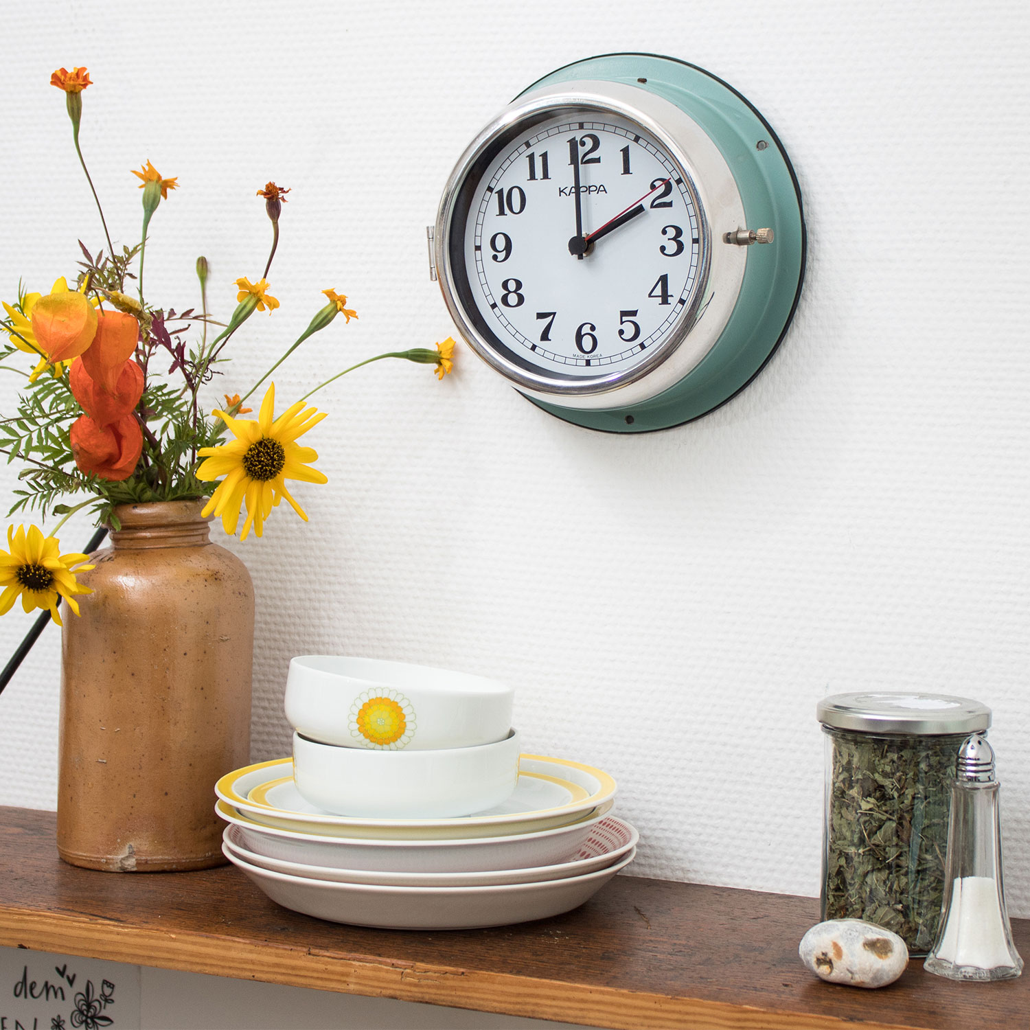 restaurierte Schiffsuhr Ein Regal aus altem Holz, darauf stehen eine hellbraune Vase mit gelben und orangen Blumen, ein Stapel Geschirr und ein Glas mit Kräutern. An der Wand hängt eine alte türkiesfarbene Schiffsuhr