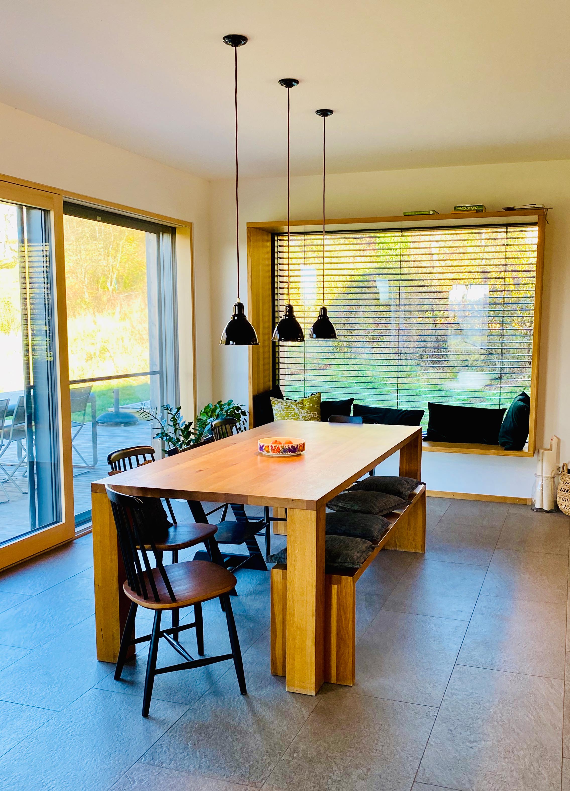 Ein moderner Wohnraum mit großen, bodentiefen Fenstern. In der Mitte steht ein großer Esstisch aus hellem Holz. Über dem Tisch hängen drei kleine, schwarze Lampen aus Emaille