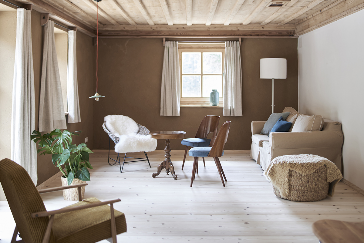 Blick in ein Wohnzimmer mit Sofa, Sesseln und alten Holzmöbeln. Von der Holzdecke hängt eine kleine weiße im Emaille Lampe.