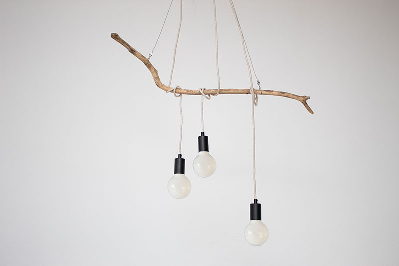 2023__Astlampe_04 Drei schwarze Lampenfassungen mit großen Glühbirnen darin, die mit ihren beigen Textilkabeln um einen alten Ast gewickelt sind, der von der Decke hängt.