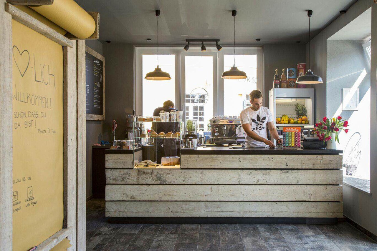 Eine Theke in einem Café. Ein Mann steht hinter der Theke und bereitet Kaffee zu. Links befindet sich auf der Theke eine Vitrine mit Kuchen, in der Mitte eine Espressomaschine. Über der Theke hängen drei Lampen aus Metall.