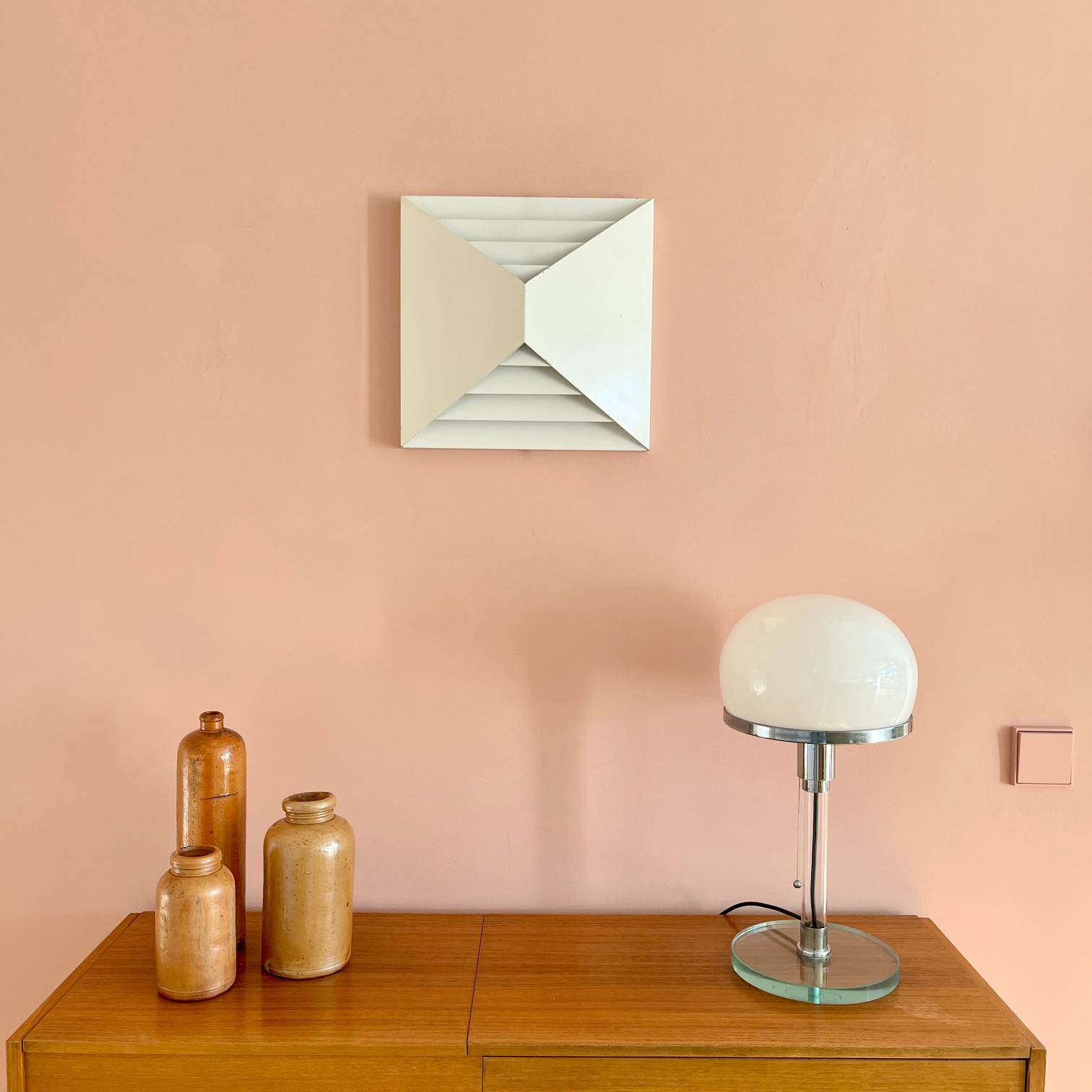 Oberfläche eines Holz-Sideboards vor einer rosa Wand. Auf dem Sideboard stehen drei Flaschen aus Steingut und eine Wagenfeld-Tischlampe. An der Wand hängt eine weiße, rechteckige Wandleuchte