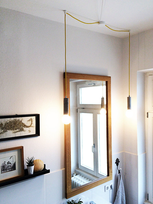 Bad Beleuchtung Blick in ein Badezimmer. Man sieht einen großen, rechteckigen Spiegel mit Holzrahmen. Links und rechts vom Spiegel hängt jeweils eine Glühbirne an einem gelben Textilkabel