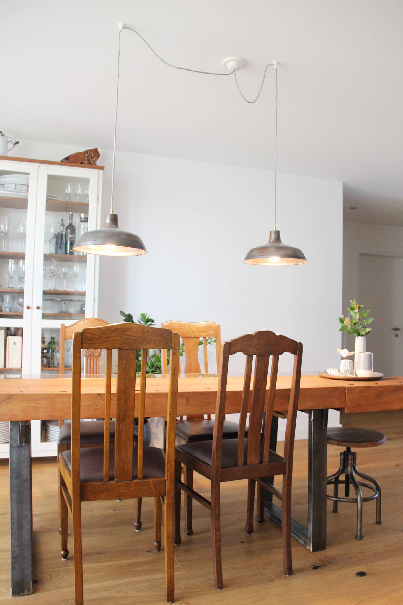 Ein Esszimmer, in der Mittevsteht ein Holztisch aus einer alten Werkbank mit vier antiken Holzstühlen darum. Über dem Tisch hängen zwei Fabrikleuchten aus Metall, die an der Decke an einem Anschluss angeschlossen sind