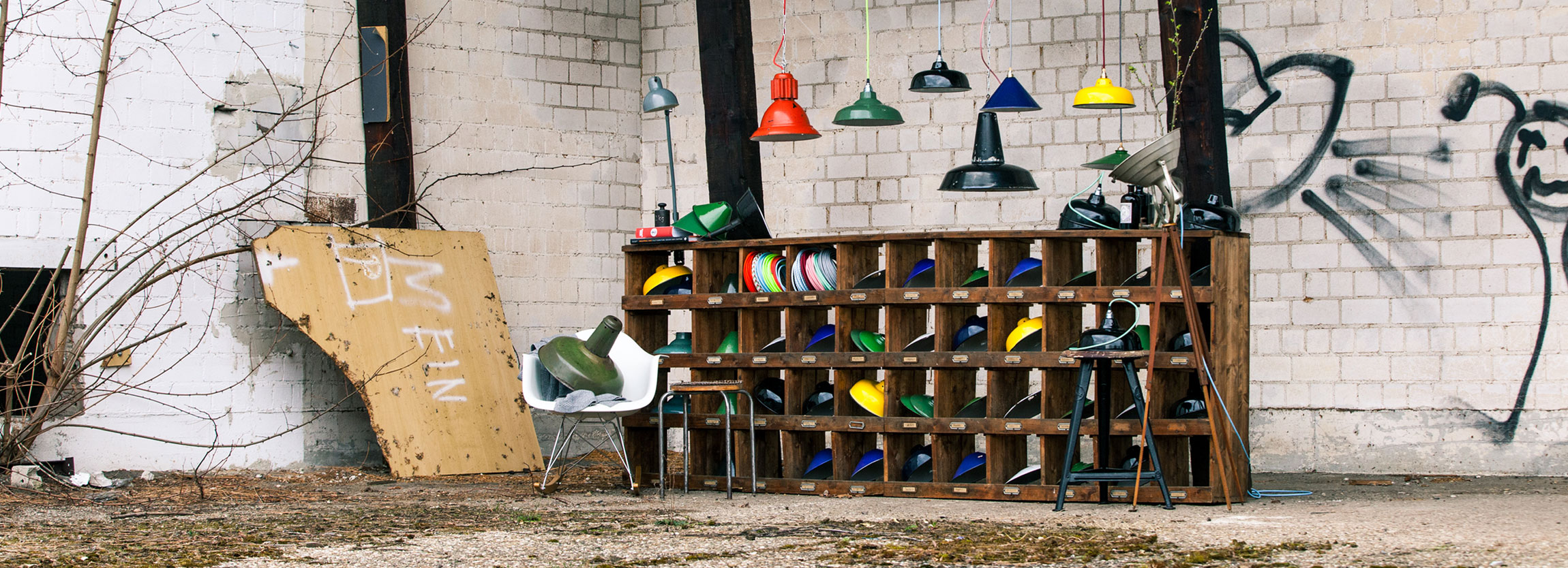 Ein verlassenes Fabrikgelände. Vor einer weißen Steinwand, die mit schwarzen Graffitis beschmiert ist, steht ein großes Holzregal mit vielen Fächern. In den Fächern sind bunte Lampenschirme und bunte Stoffkabel. Über dem Regal hängen alte Fabriklampen in 